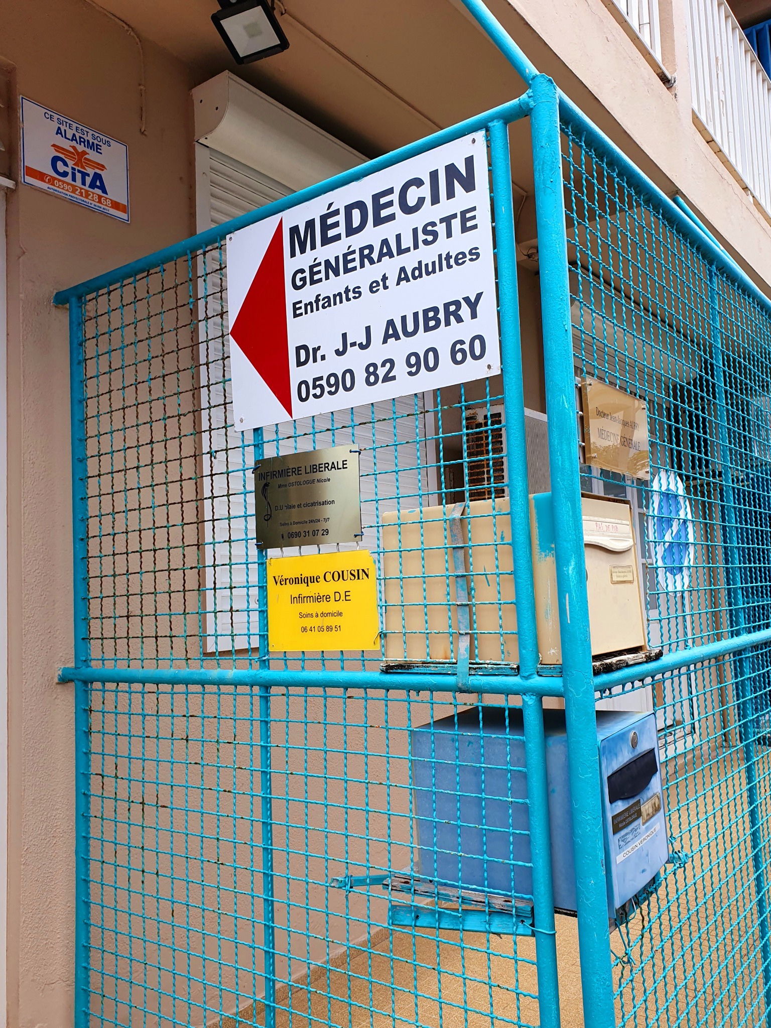 Panneau d'accès « Médecin généraliste, enfants et adultes, Dr J-J Aubry » fixé sur la grille bleue